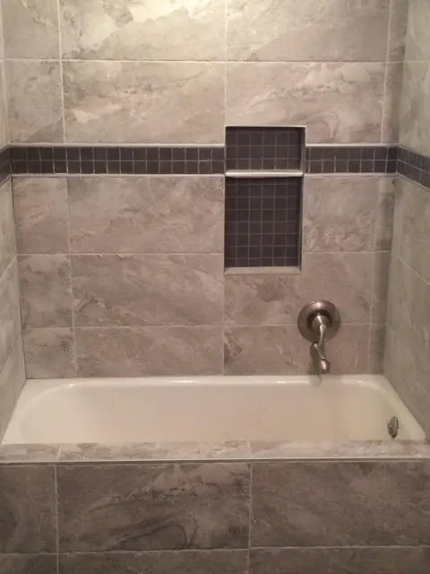 Tub shower surround in gray stone-look tile with dark mosaic accent band and niche