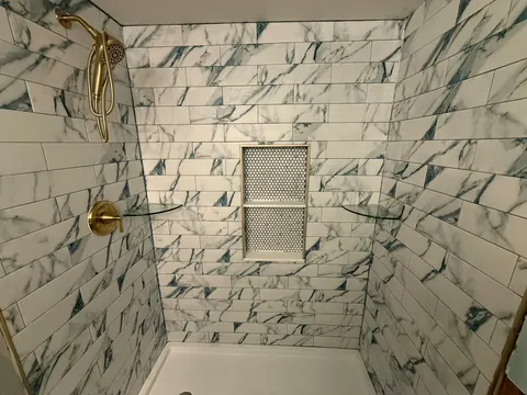 Marble-look subway tile shower with brushed gold hand shower and valve, glass corner shelves and a penny tile niche