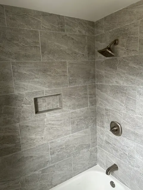 Completed tub shower surround in gray stone-look tile with brushed nickel fixtures and mosaic niche