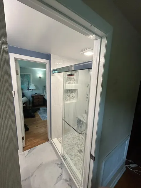 View from the hall into a new walk-in shower with a sliding glass door and marble-look porcelain floor