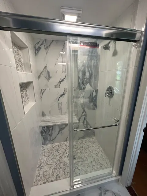 Finished walk-in shower behind a sliding glass door, with pebble mosaic floor, tiled bench and recessed niches