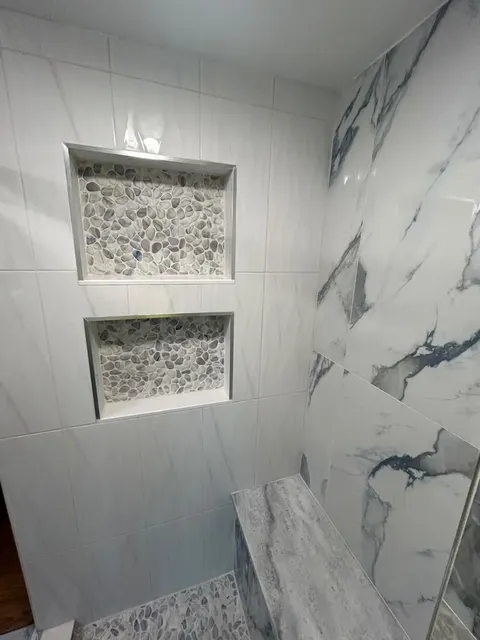Two stacked shower niches with pebble mosaic backs and metal edge trim beside a stone-topped shower bench